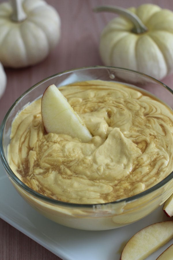 Bowl of pumpkin cheesecake dip topped with caramel swirl and an apple slice for dipping, surrounded by white pumpkins