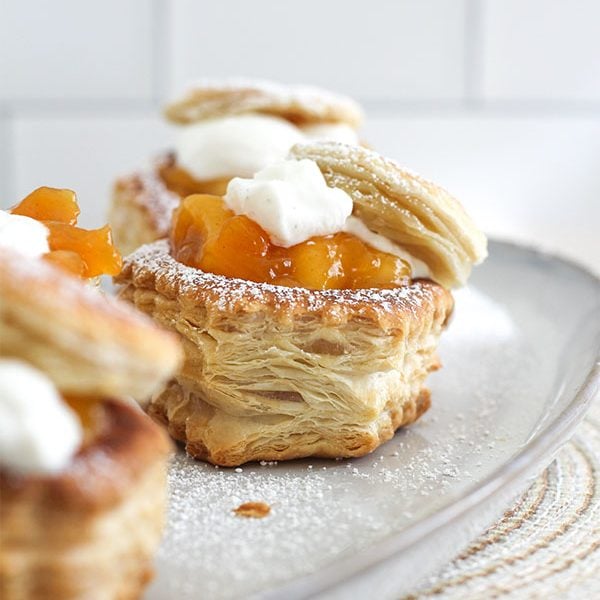 Close-up of peach pie pastry cups on a serving plate, filled with peach chunks, topped with whipped cream and a dusting of powdered sugar