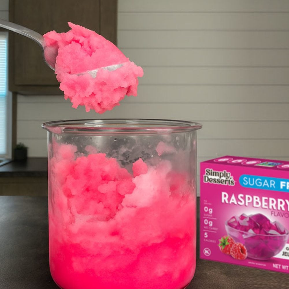 Ninja Creami Pint with pink raspberry lemonade sorbet, with a spoonful lifted above the cup and a Simply Desserts Raspberry Jel mix box in the background on a kitchen counter