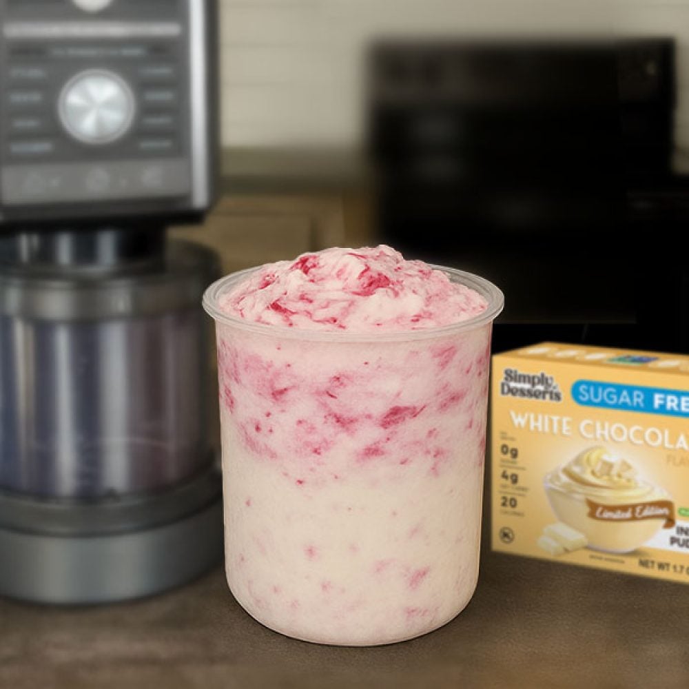 Freshly blended white chocolate raspberry cheesecake ice cream in a Ninja Creami pint container beside a Simply Desserts White Chocolate Pudding Mix box